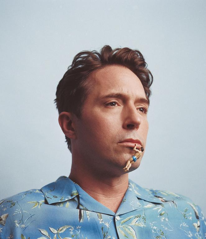 A man in a blue patterned shirt has a small figurine of a swimmer hanging from his lower lip.
