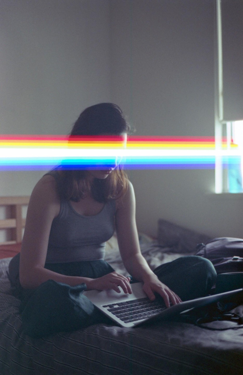 A woman sits on a bed using a laptop, with a rainbow light streak across her face.