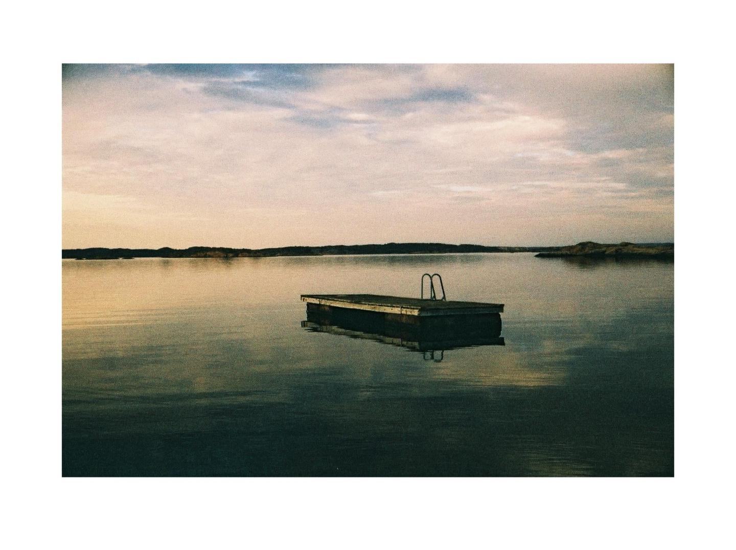 A wooden swimming platform with a ladder floats on a calm lake under a cloudy sky.