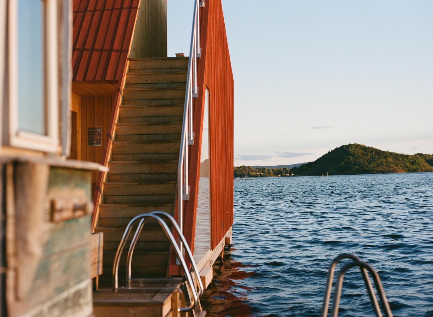 A red wooden building with stairs leading into the water is shown next to a body of water with a forested hill in the distance.