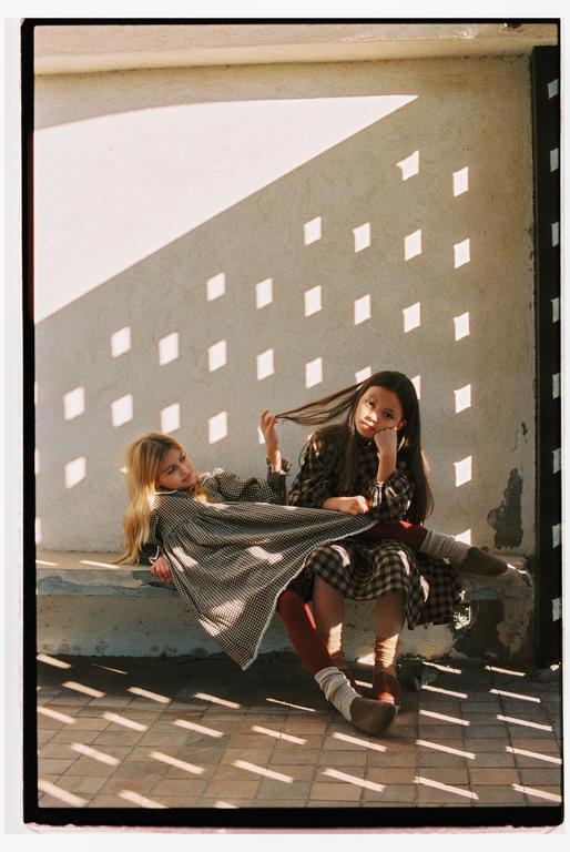 Two girls sit on a concrete bench with diamond-shaped shadows cast on the wall behind them.
