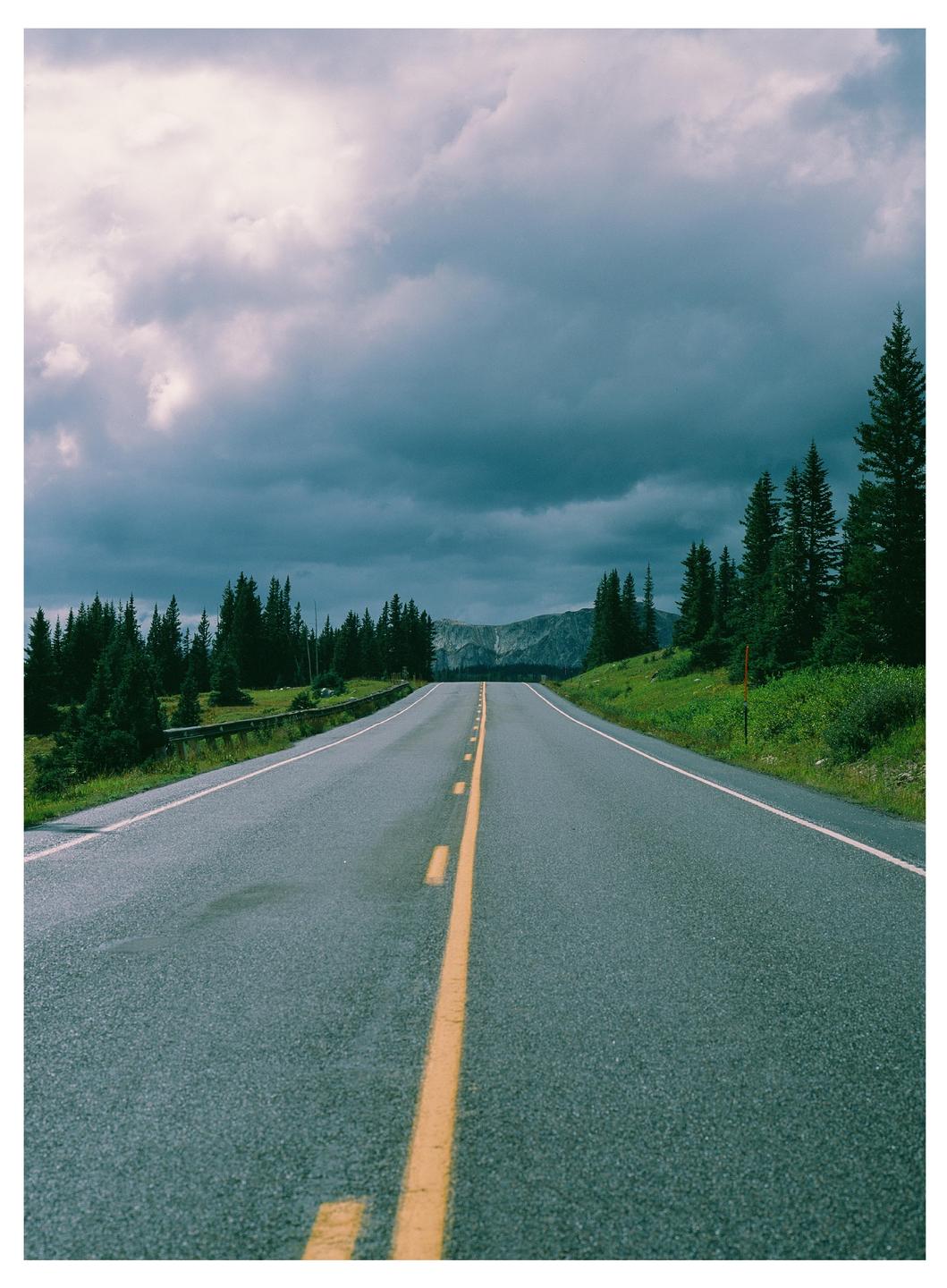 A long road with a yellow dividing line leads toward distant mountains under a cloudy sky.