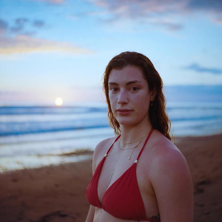 A woman in a red bikini stands on a beach with the ocean and sunset in the background.