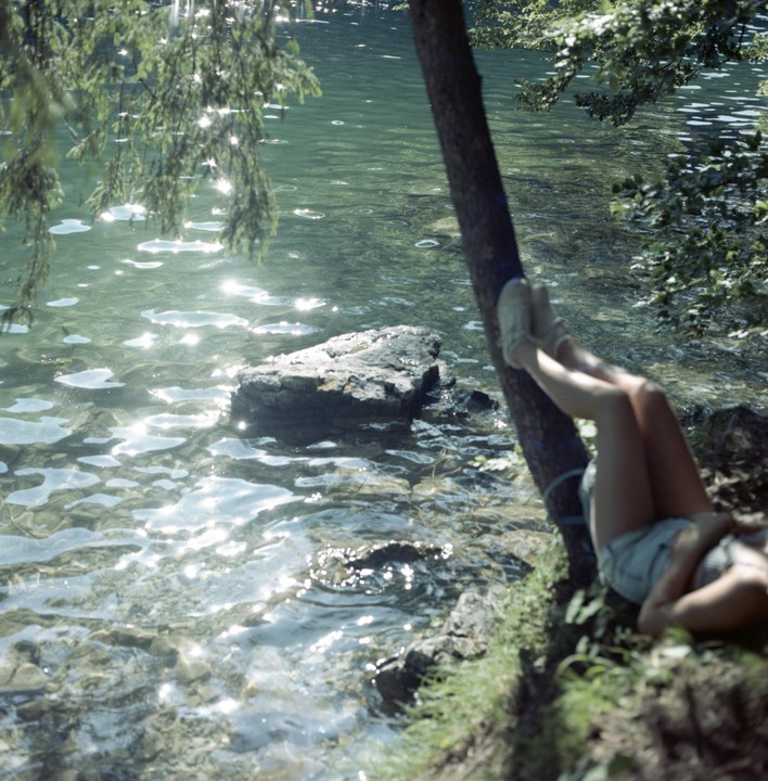 A woman lies on the bank of a lake with her legs resting against a tree.