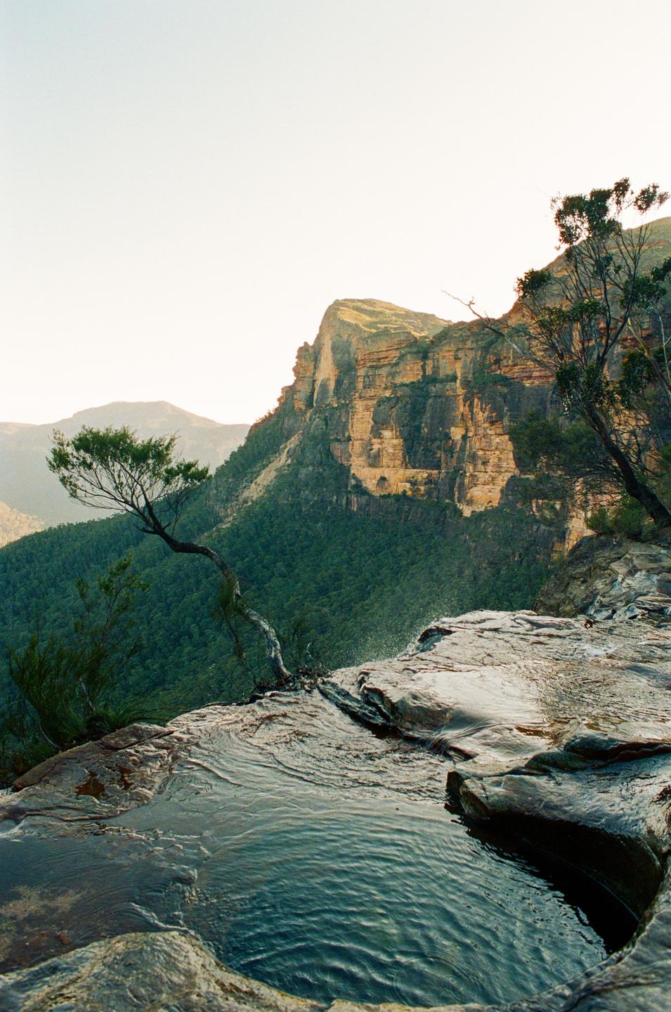 A small pool of water on a rocky cliff edge overlooks a vast valley with mountains and trees.