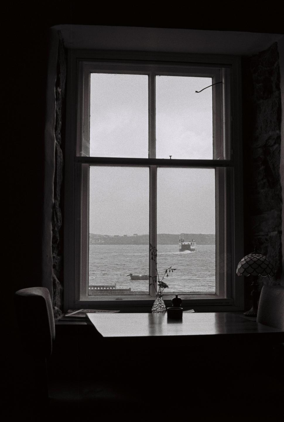 A ferry boat crosses a body of water, viewed from a window with a table and lamp in the foreground.