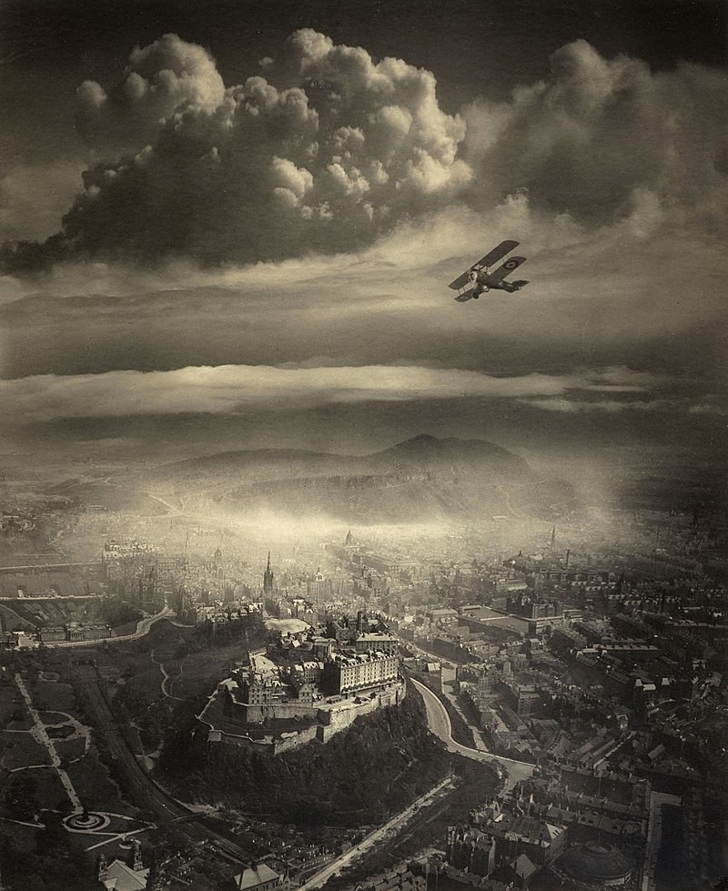An aerial view shows a biplane flying over Edinburgh Castle and the city, with mountains in the hazy distance under a cloudy sky.