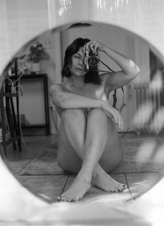 A woman sits on the floor, holding a camera to her face, reflected in a mirror.