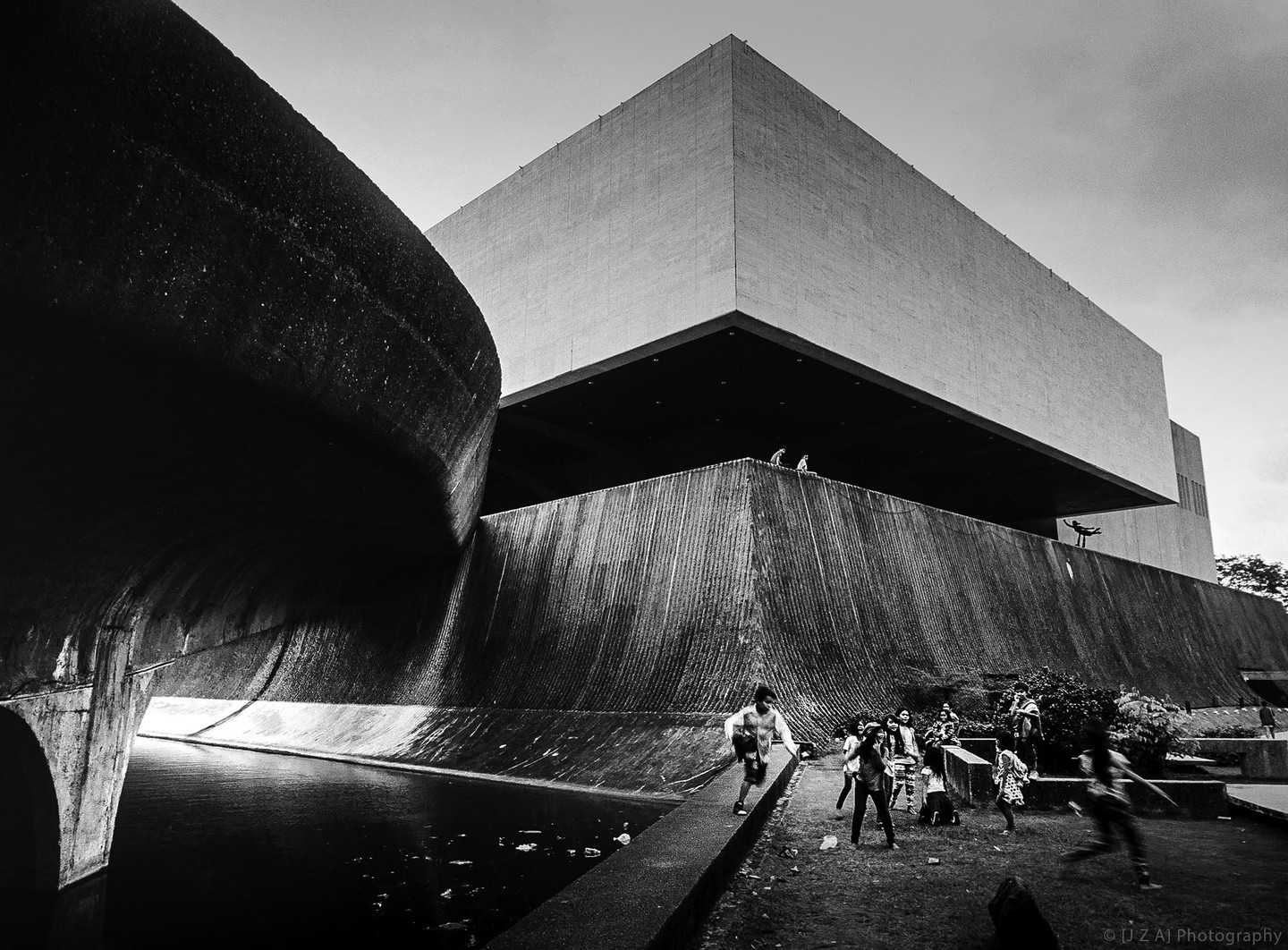 Children play on a grassy area next to a body of water and a large brutalist building.