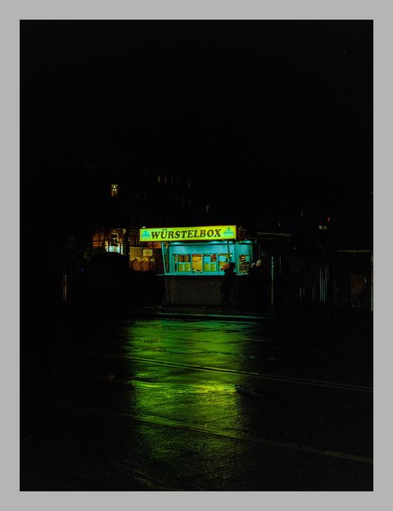 A brightly lit food stand with a green sign reading "WÜRSTELBOX" reflects onto a wet street at night.