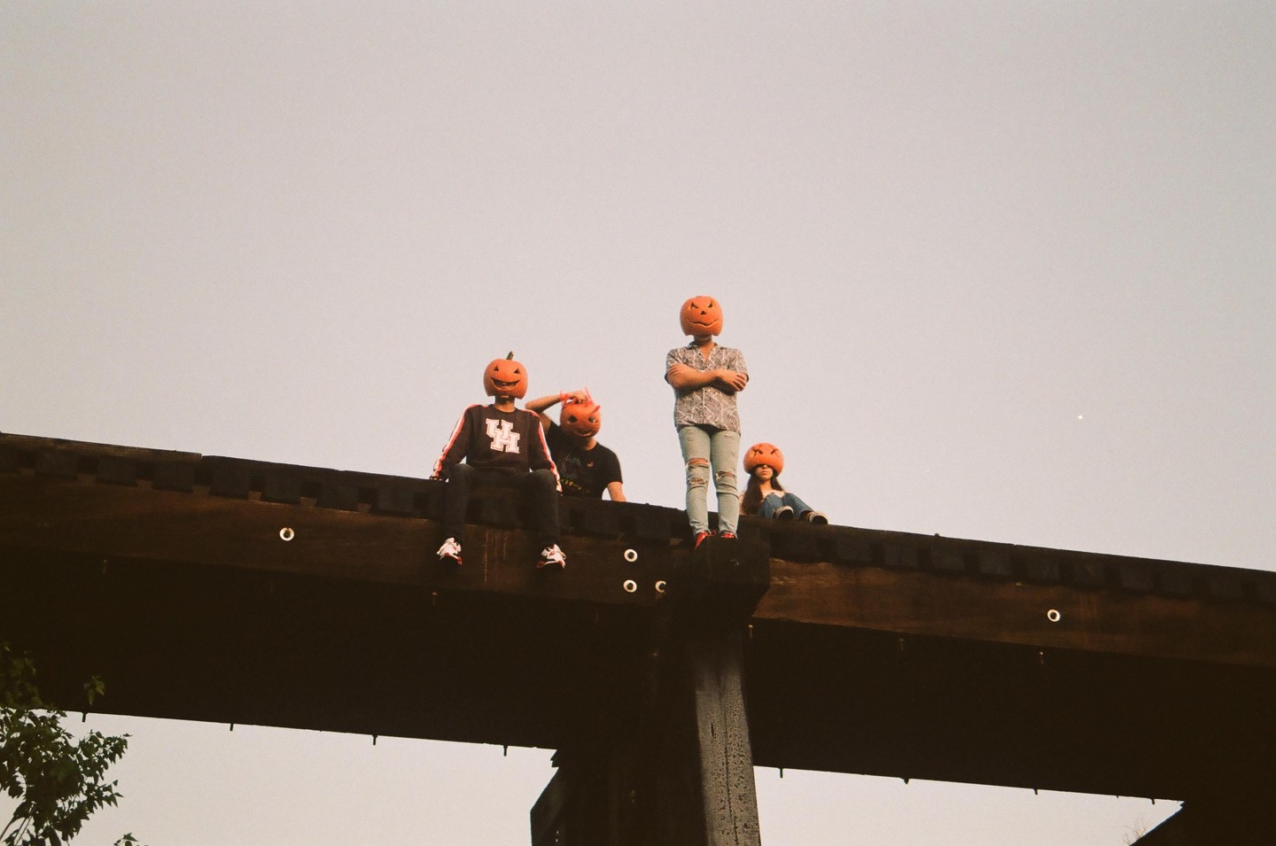 Four people wearing pumpkin masks are sitting and standing on a wooden bridge.