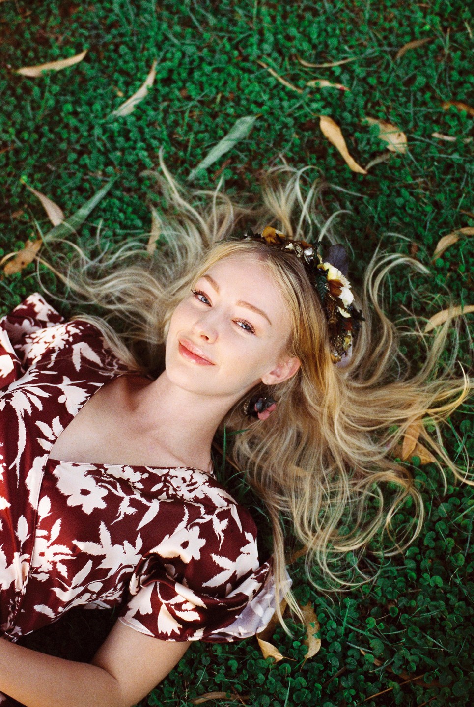 A woman with long blonde hair and a floral headband lies on green grass with fallen leaves.