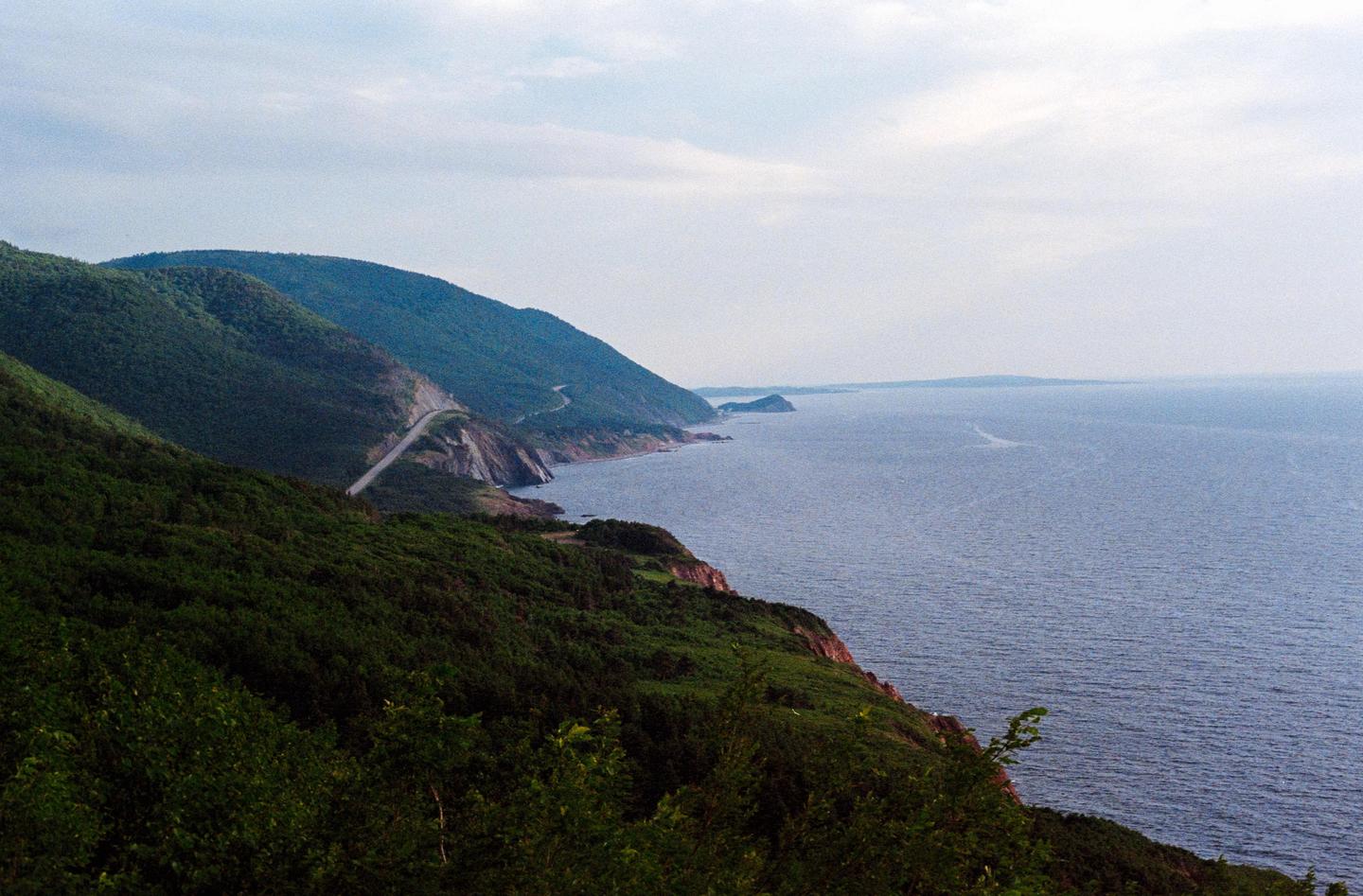 A winding road follows the coastline along a forested mountain range next to a large body of water.
