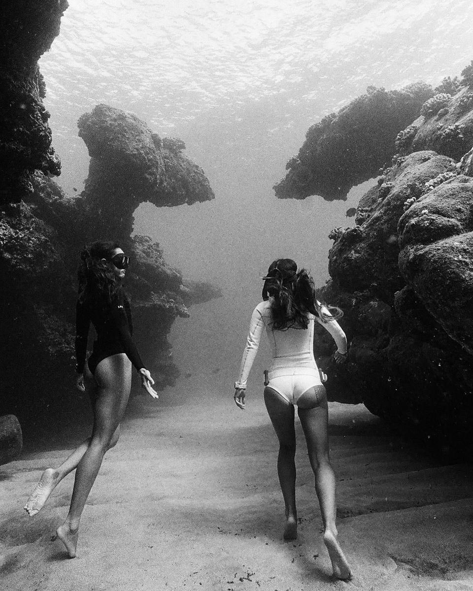 Two women in swimsuits and goggles swim underwater between rock formations.
