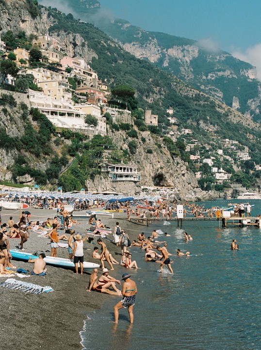 A crowded beach with people swimming and sunbathing is nestled at the base of a steep, building-covered hillside.