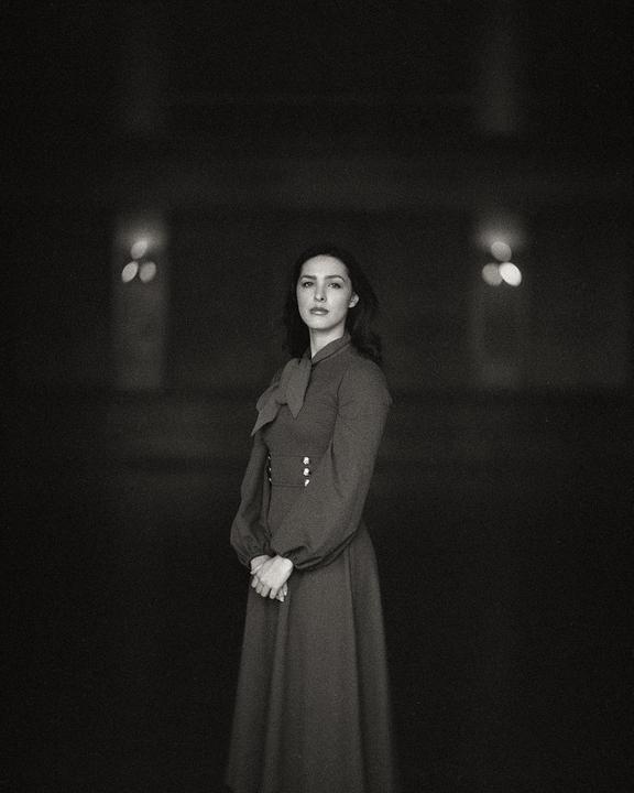 A woman in a long dress stands with her hands clasped in front of her, looking directly at the viewer.