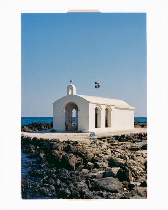 A small white church with a bell tower and a flag stands on a rocky shore next to the sea under a clear blue sky.