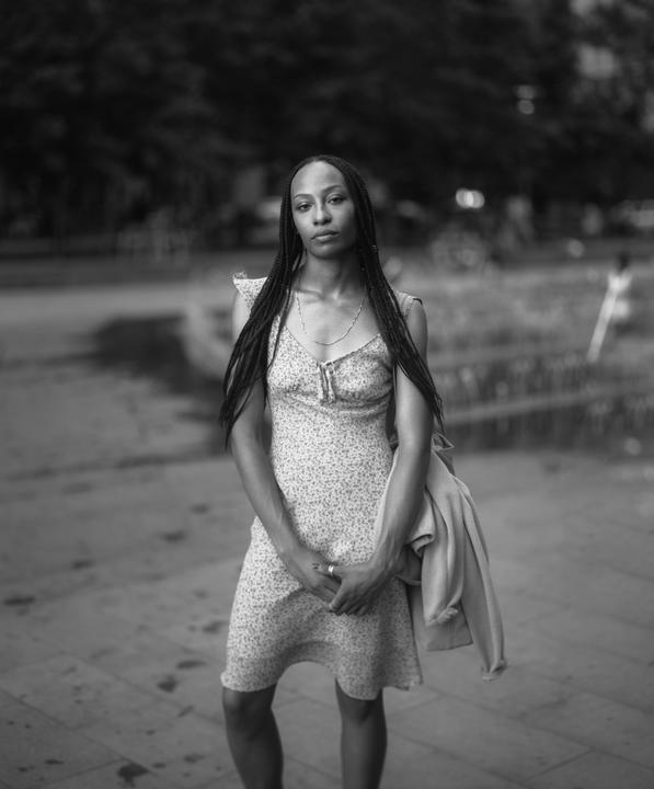 A woman with long braids and a floral dress stands outdoors with a jacket draped over her arm.