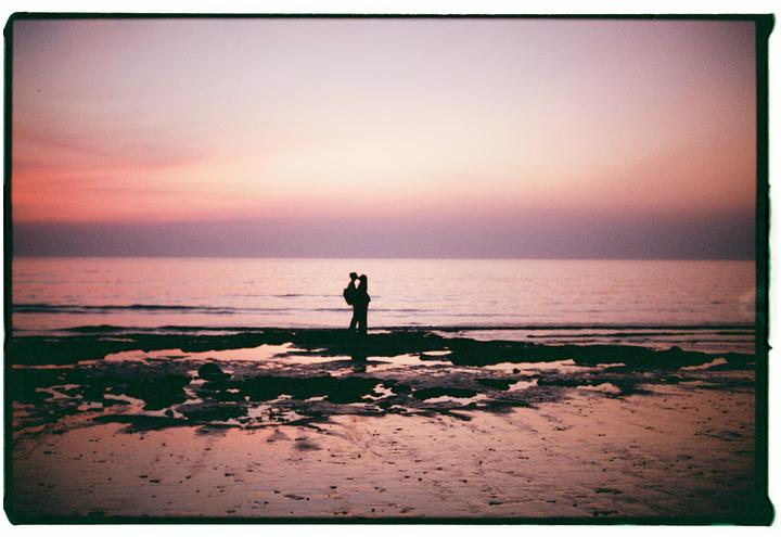 A couple stands silhouetted against the ocean at sunset on a beach.