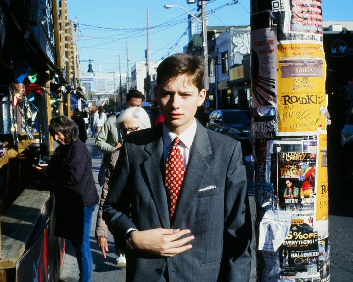 A young man in a suit and red tie stands on a sunny street next to a pole covered in posters.