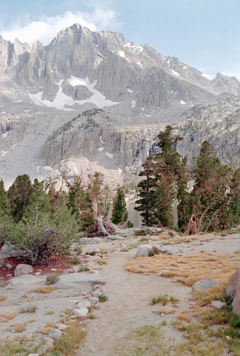 A dirt path leads through a rocky, forested area towards a lake and snow-capped mountains.