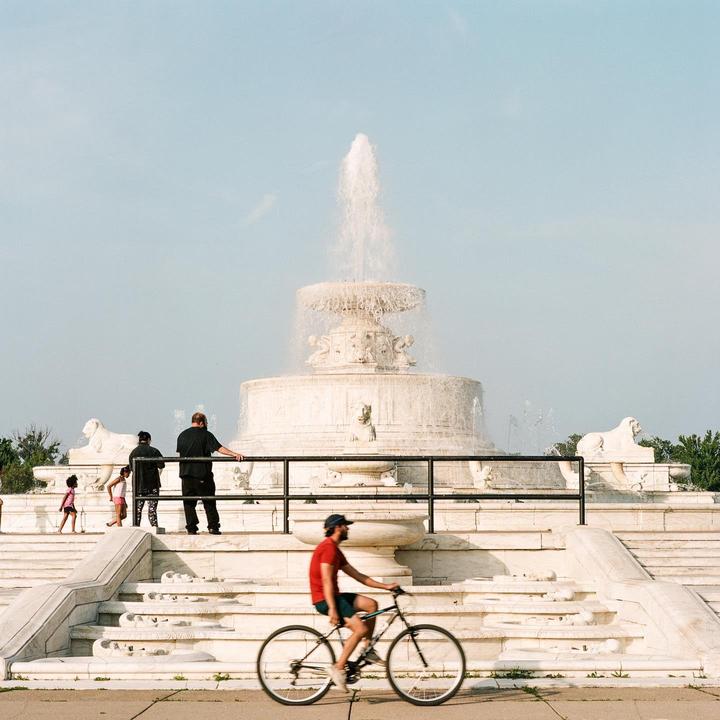 A man on a bicycle rides past a large white fountain with people standing on its steps.