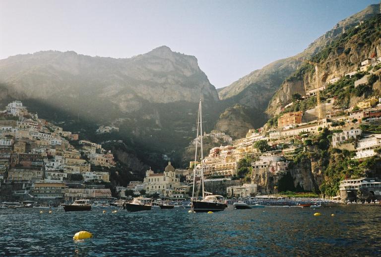 A coastal town with buildings climbing up a steep mountain is seen from the water, with boats moored in the foreground.