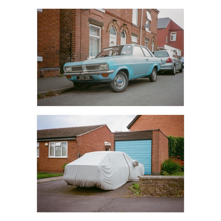 A light blue car is parked on a street next to a brick building, and another car is covered with a tarp in a driveway.