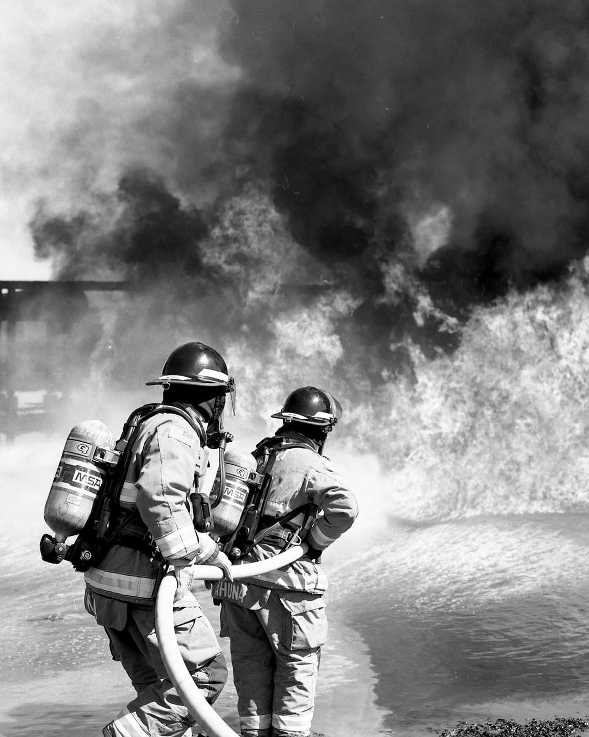 Two firefighters spray water on a large fire with thick smoke.