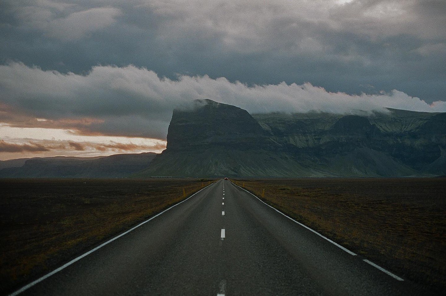 A long, straight road leads towards a large mountain partially obscured by clouds under a dramatic sky.