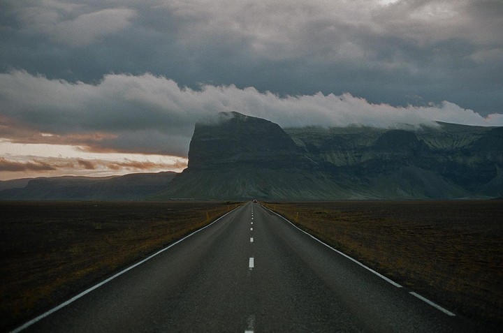 A long, straight road leads towards a large mountain partially obscured by clouds under a dramatic sky.