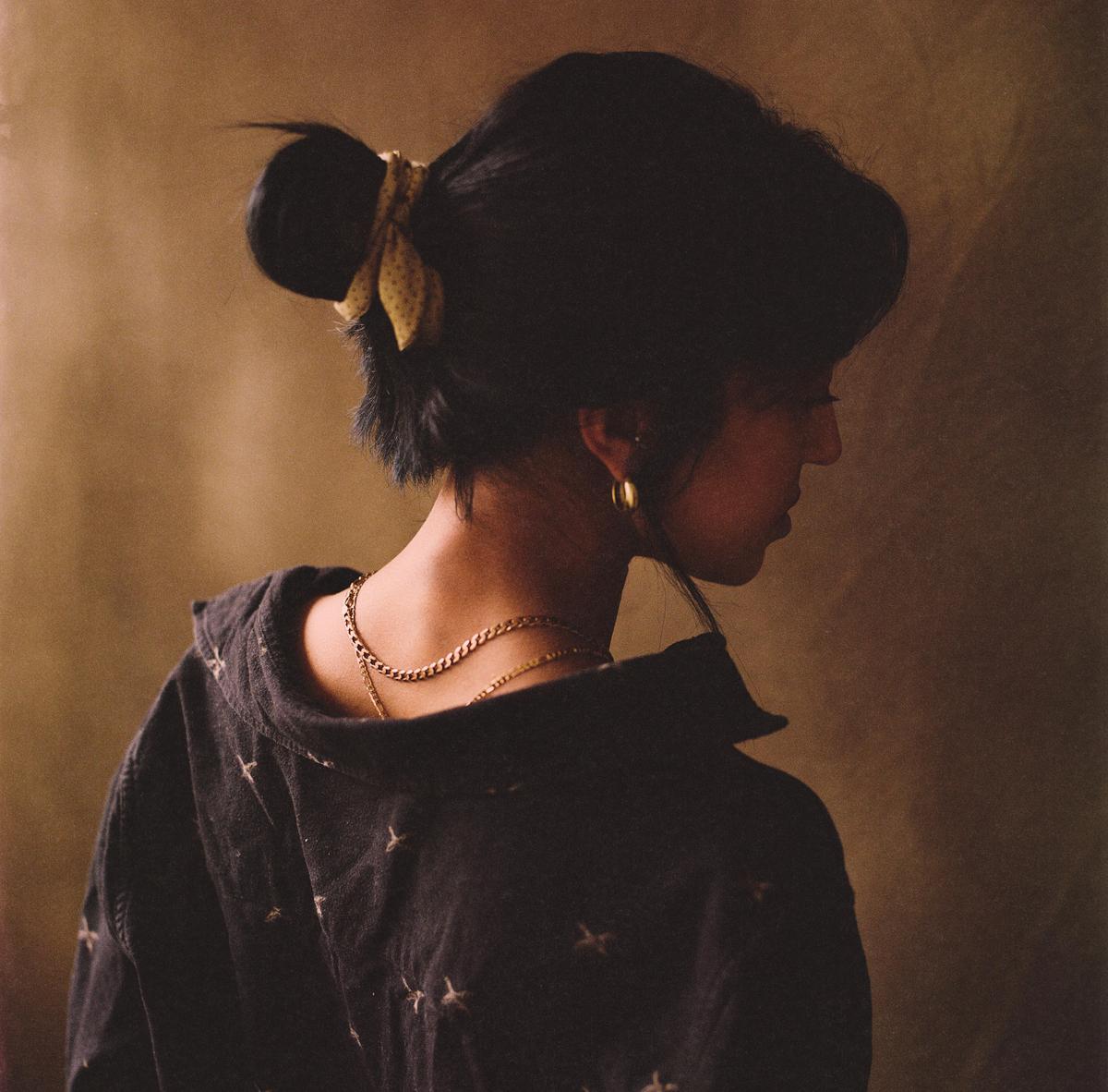 A woman with dark hair tied in a bun with a yellow ribbon is seen from behind, wearing a dark shirt and gold necklaces.