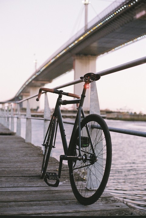 A bicycle with a red tail light is parked on a wooden pier next to a body of water, with a large bridge illuminated by lights in the background.