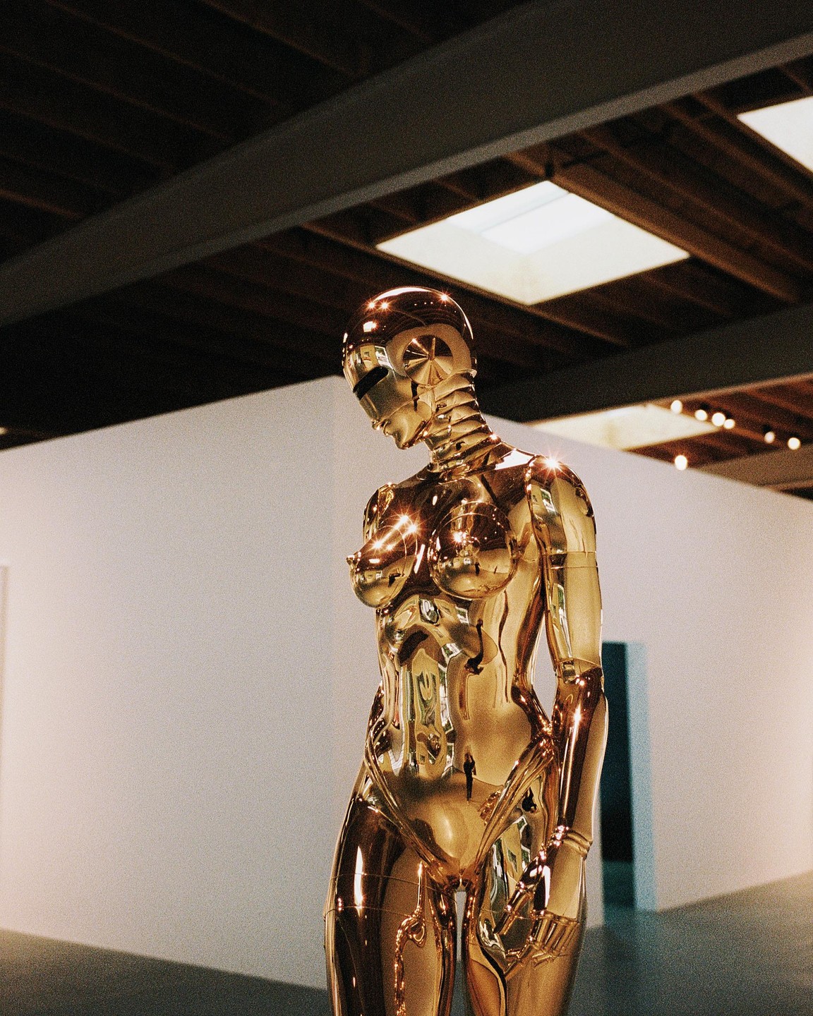 A golden, shiny, humanoid robot stands in a room with a white wall and a dark ceiling.