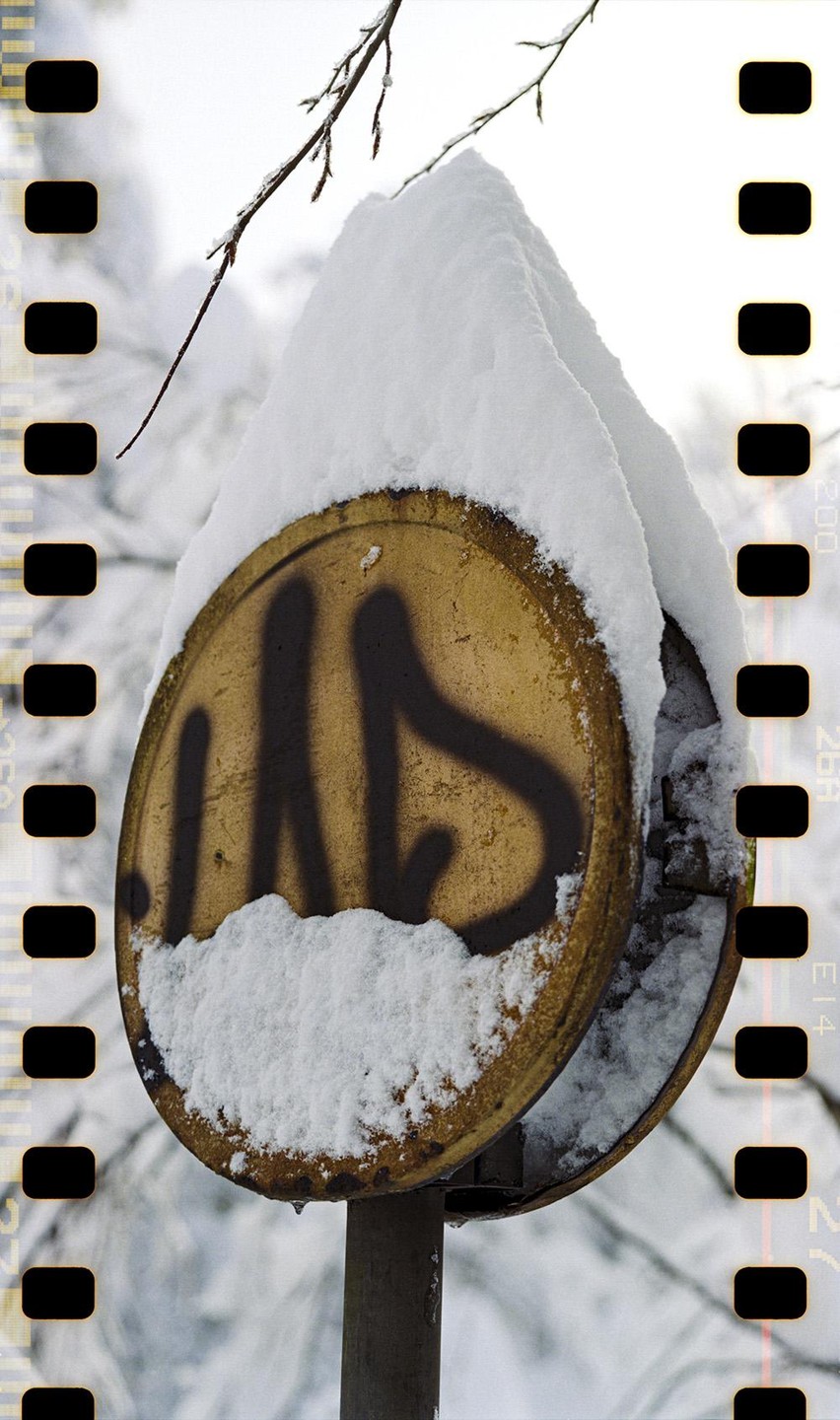 A round, yellow sign with black graffiti is covered in snow, with a bare branch visible above it.