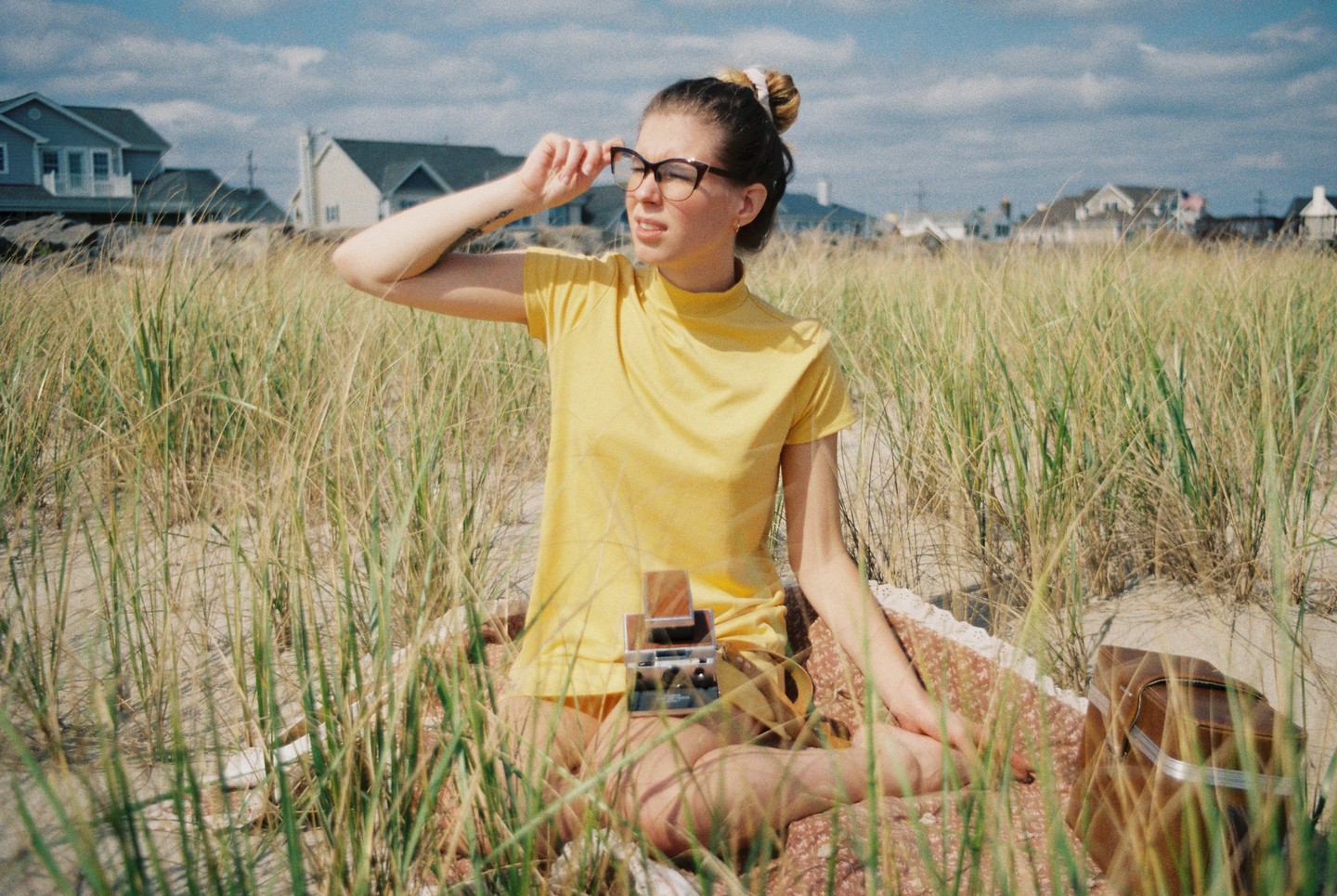 A woman in a yellow shirt sits on a blanket in tall grass, adjusting her glasses.