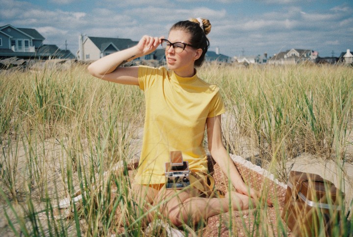 A woman in a yellow shirt sits on a blanket in tall grass, adjusting her glasses.