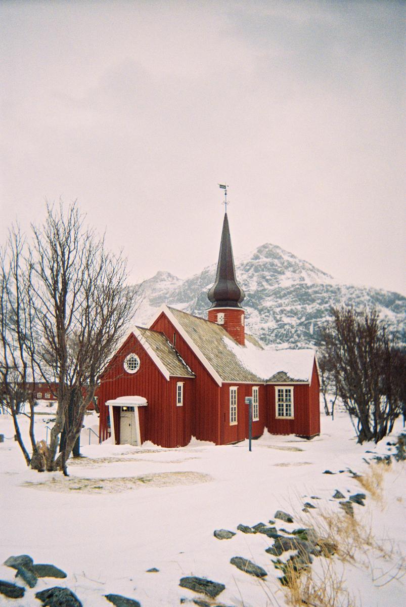 A red church with a steeple stands in a snowy landscape with mountains in the background.
