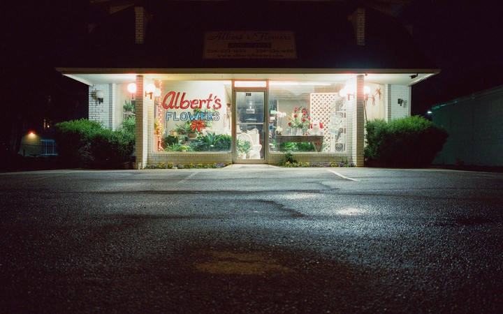 A brightly lit flower shop with a sign that reads "Albert's Flowers" stands at night.