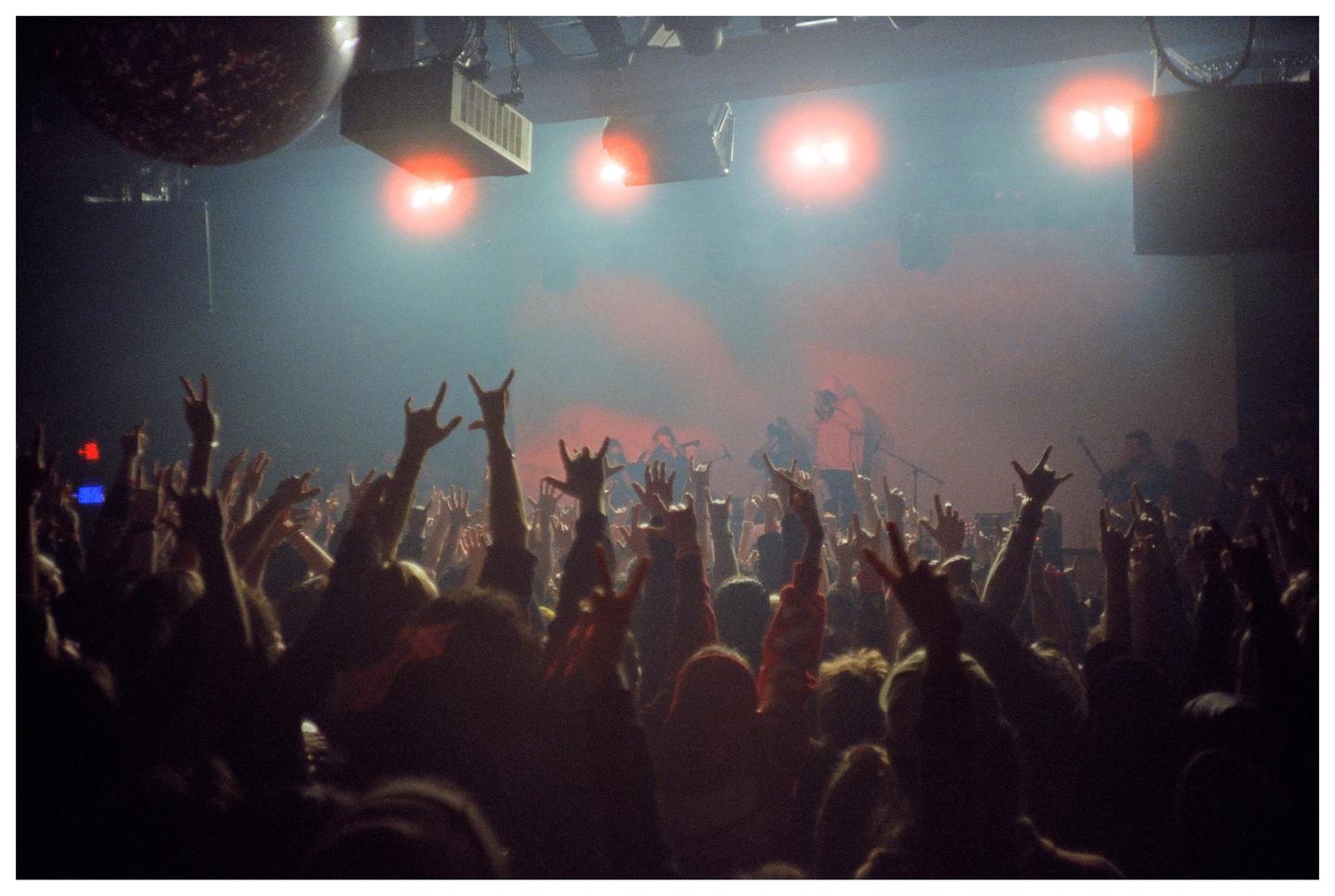 A crowd of people with their hands raised in a concert venue are illuminated by red stage lights.