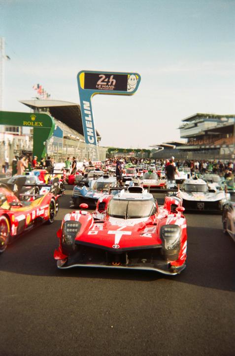 A red race car is in the foreground of a line of race cars on a track with a "24h Le Mans" sign in the background.