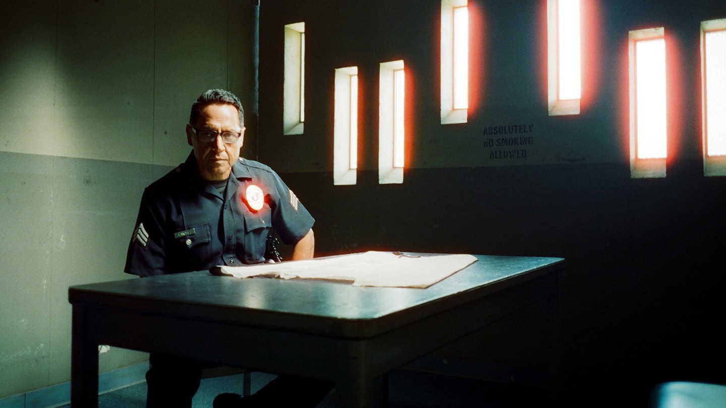 A man in a police uniform sits at a table with papers on it, with a wall behind him that has several rectangular windows and the words "ABSOLUTELY NO SMOKING ALLOWED" stenciled on it.