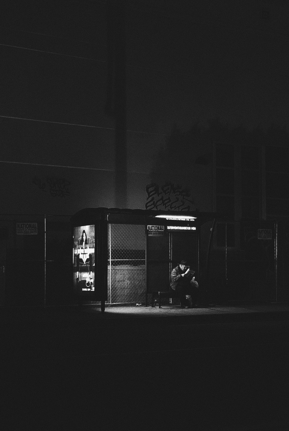 A man sits hunched over on a bench inside a bus stop at night, illuminated by a light from within the shelter.