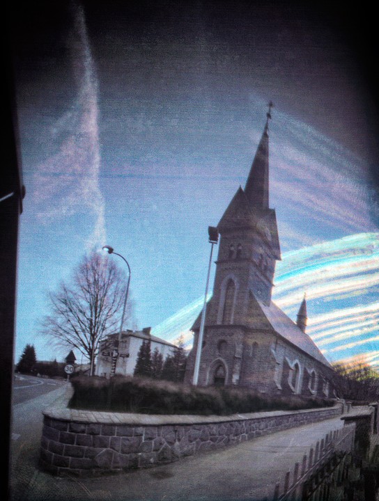 A church with a tall steeple stands beside a road with a stone wall in the foreground under a streaky sky.