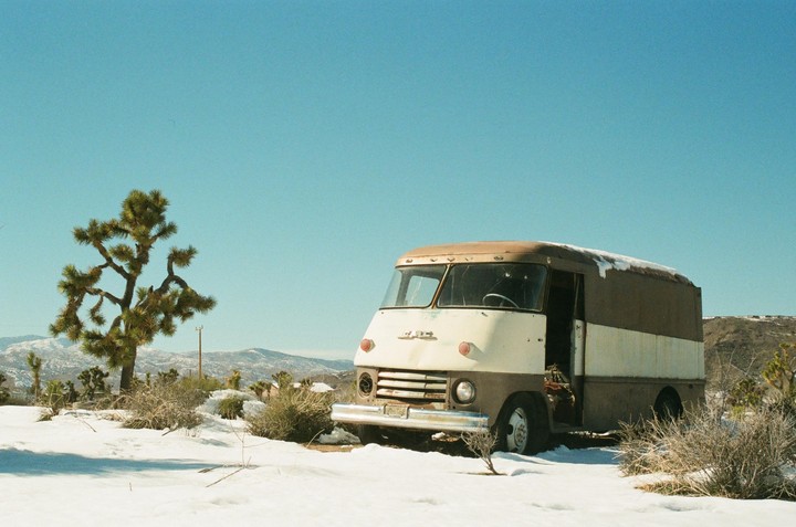 A brown and white van is parked in a snowy desert landscape with a Joshua tree and mountains in the background.