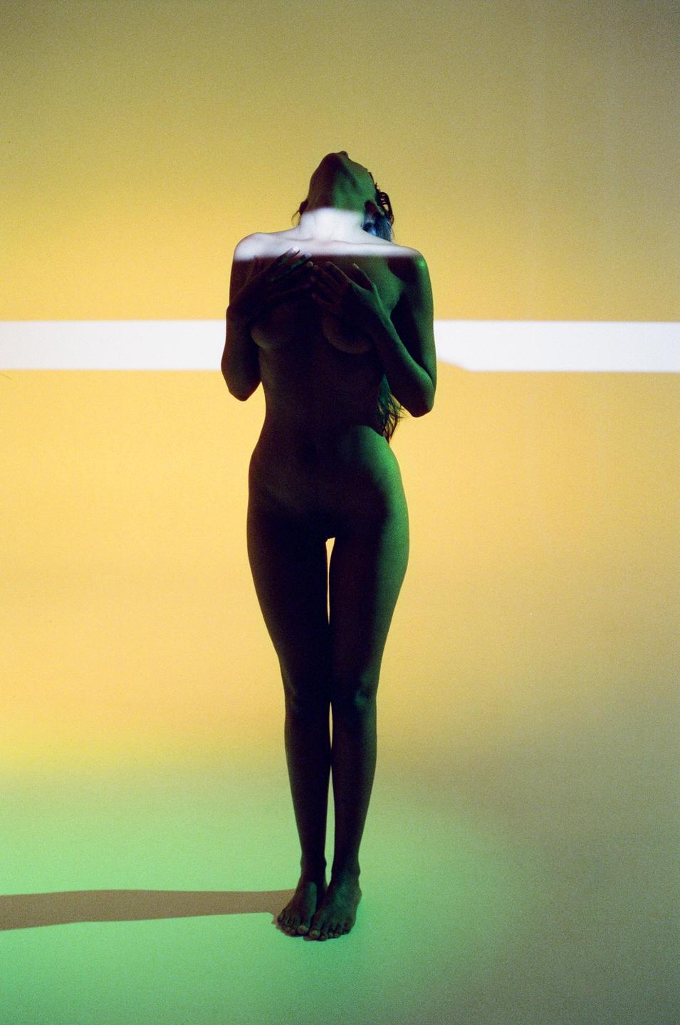 A nude woman stands with her head tilted back and hands covering her chest, illuminated by green and yellow light.