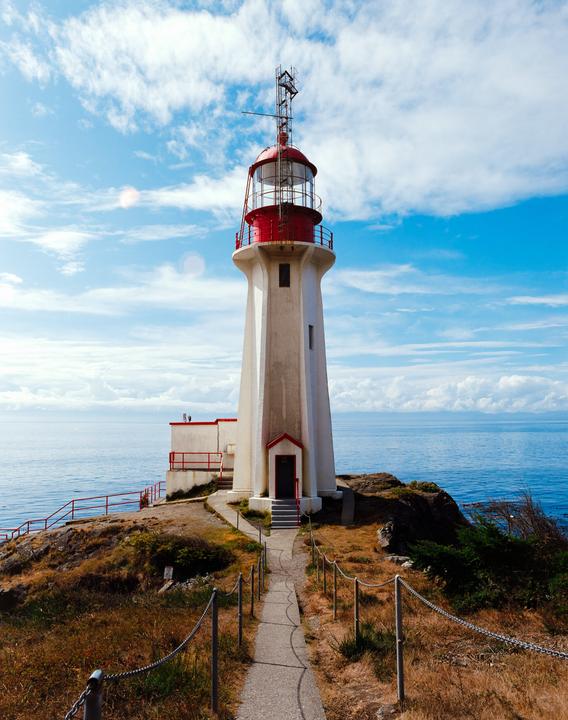 A white lighthouse with a red top stands on a rocky cliff overlooking the ocean under a blue sky with white clouds.