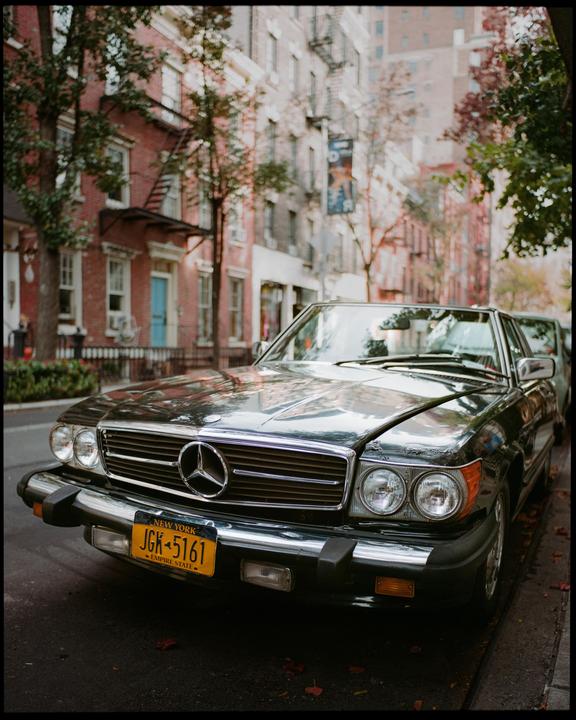 A black car with a New York license plate is parked on a street lined with brick buildings and trees.