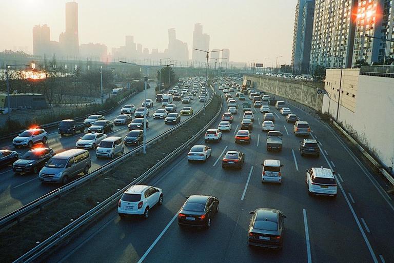 A multi-lane highway is filled with cars during golden hour, with a city skyline in the hazy distance.