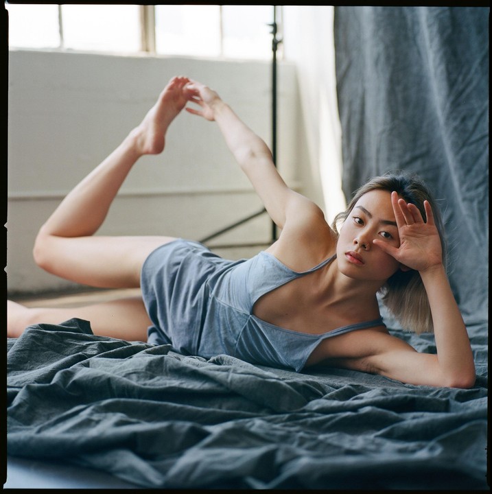 A woman in a blue dress is lying on a gray sheet with one leg bent and her foot raised.